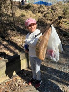 A young girl with a pink hat holds up a litter grabber with several bags of trash on it